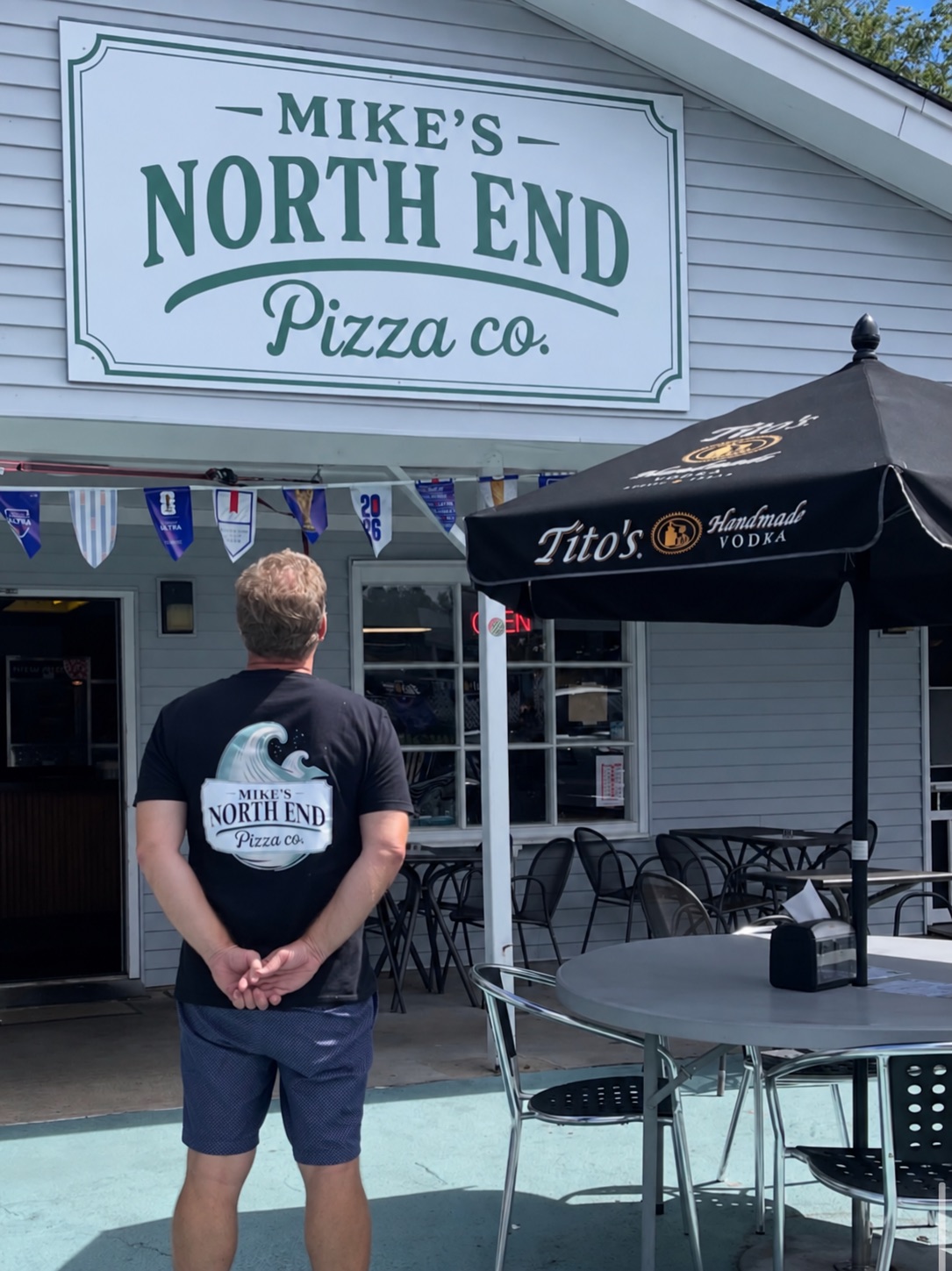 A man standing outside Mike's North End Pizza Co. in a wave-logo shirt, looking up at the shop sign